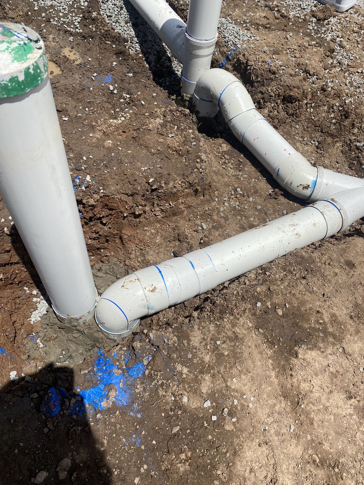 White PVC plumbing pipes are connected in a trench, leading to a vertical pipe with a green cap in the dirt.