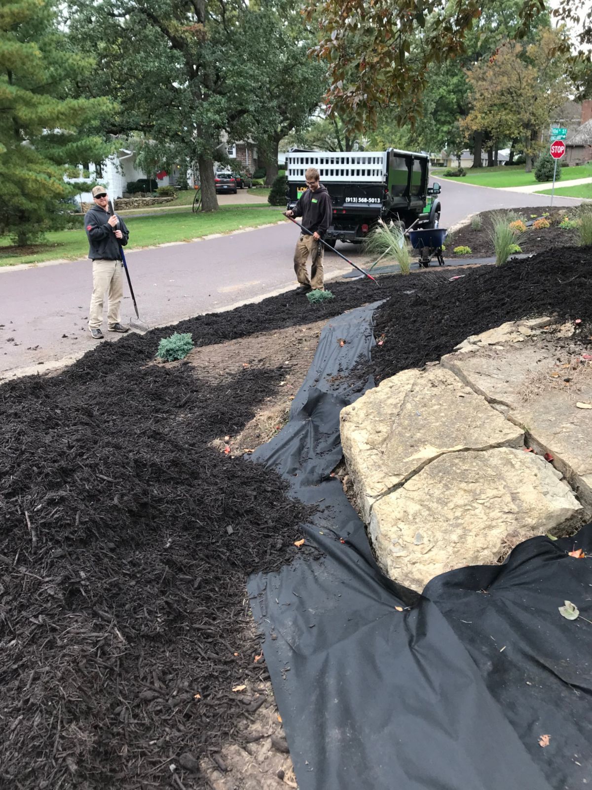 Two workers spread black mulch over landscape fabric in a residential front yard near a large stone and a utility vehicle.