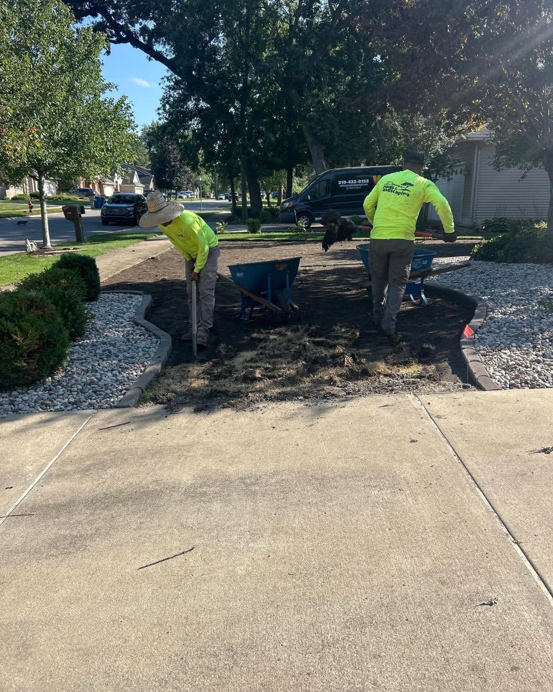 Two workers in bright green shirts move soil in a residential front yard, using shovels and a wheelbarrow.
