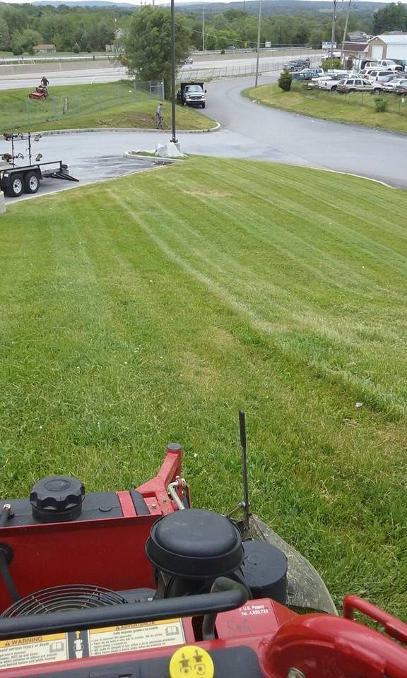 First-person view from a red riding lawn mower cutting a green grass slope near a road, parking lot, and utility pole.