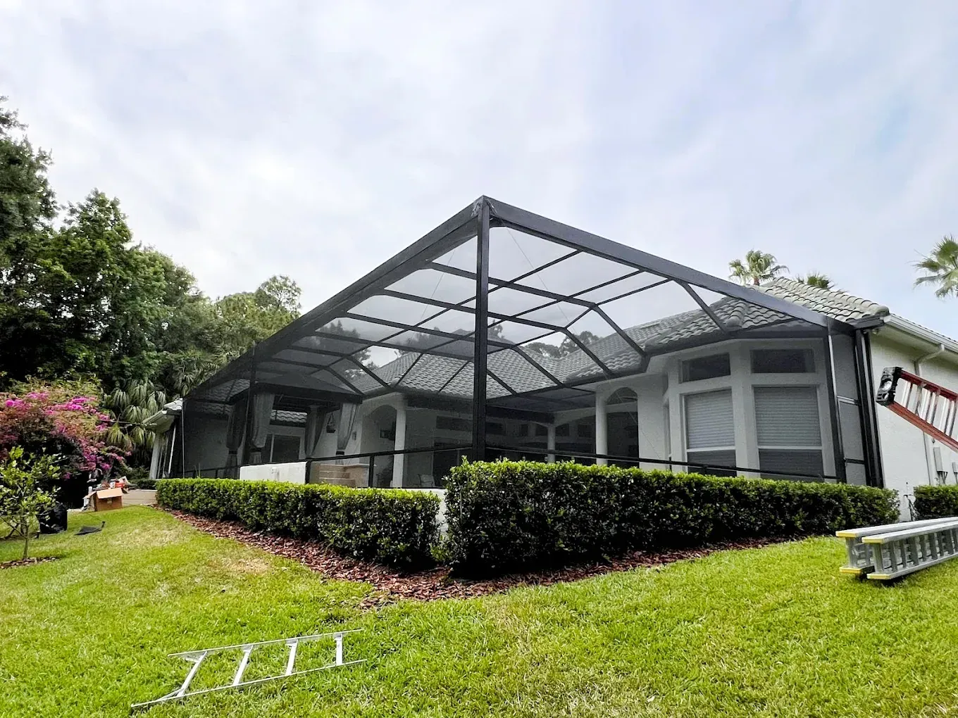 A screened-in patio with a dark frame, attached to a white house. Green lawn, bushes, and trees surround.