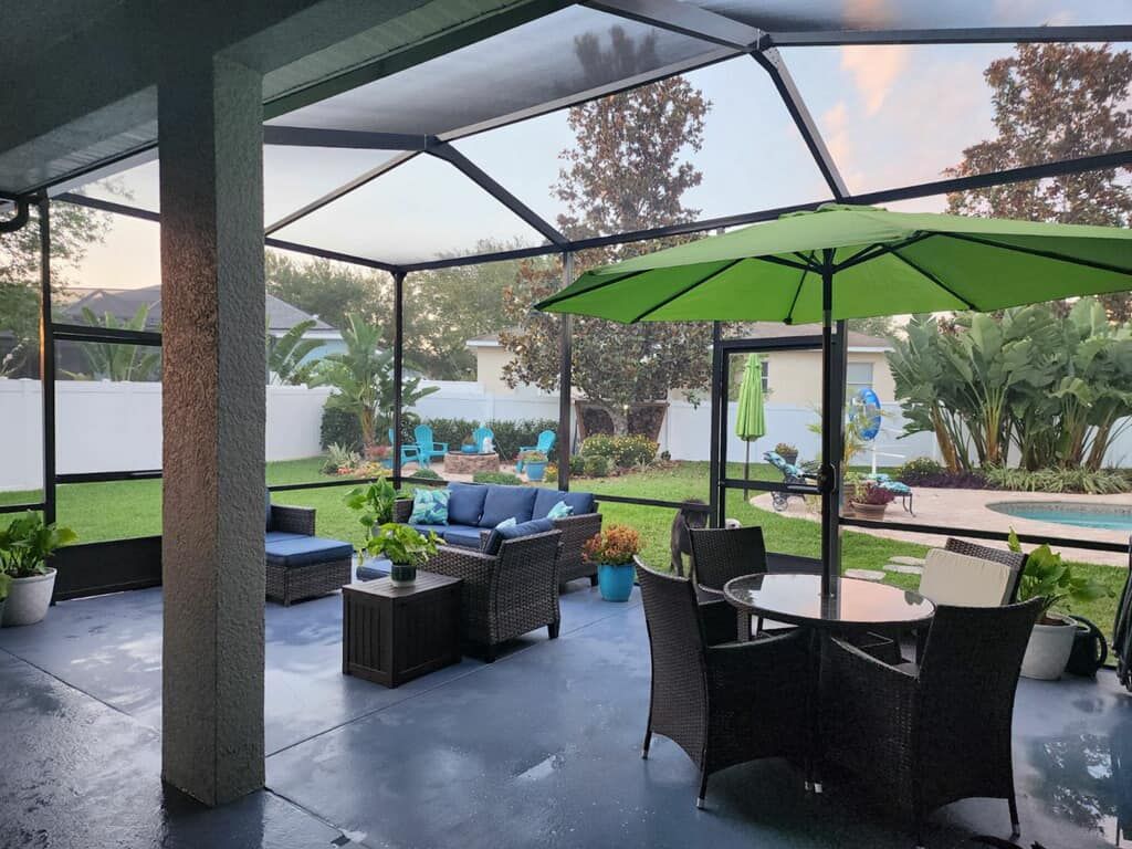 Screened-in patio with outdoor furniture: couches, table, and umbrella. Green lawn and a pool in the background.