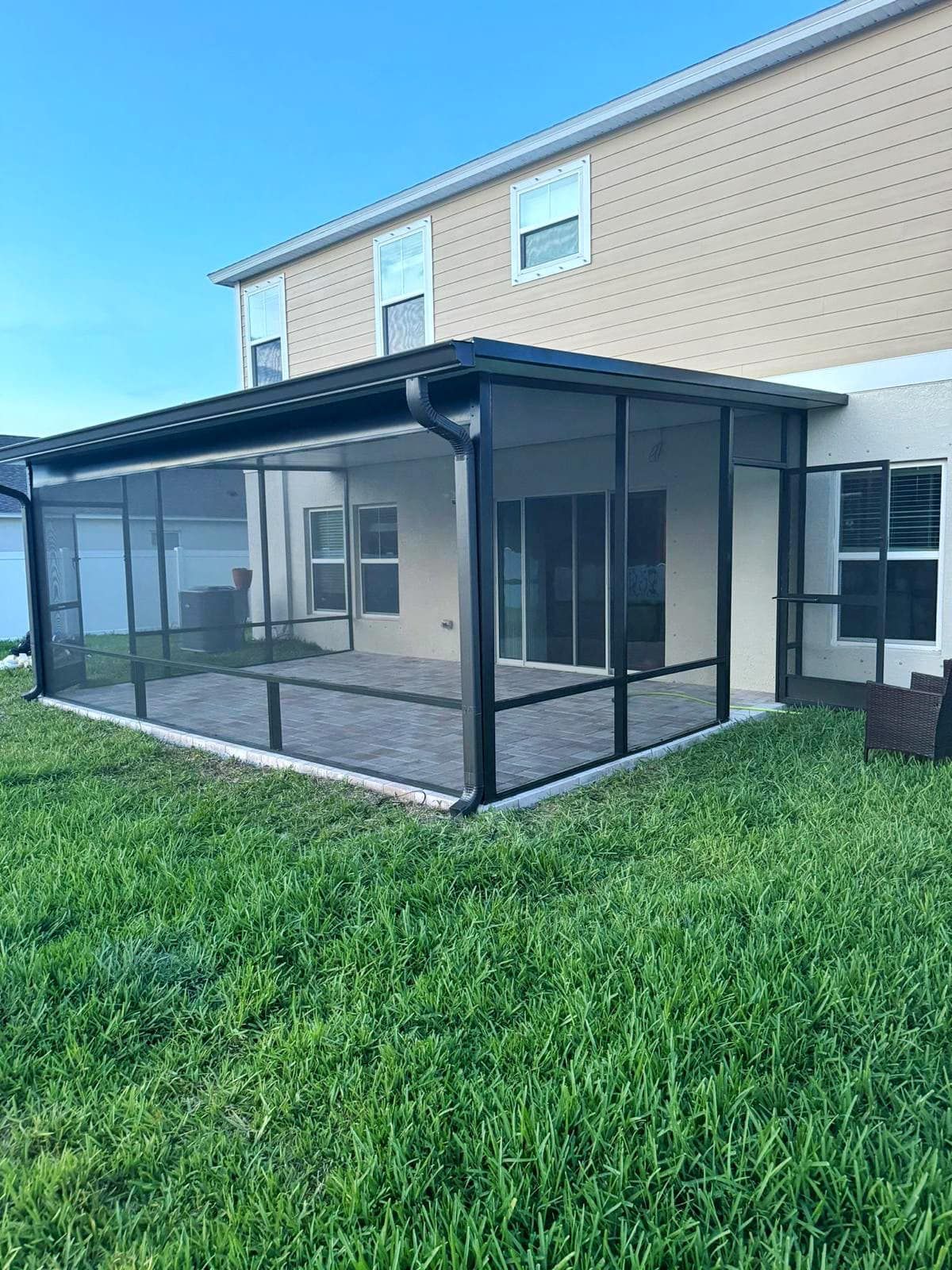 Screened-in patio attached to a light beige house with dark trim, located over a brick patio and surrounded by green grass.