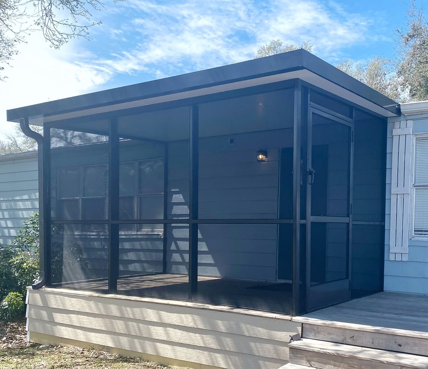 Screened porch with black frames, dark screens, and a light-colored roof, attached to a house with a wooden deck.