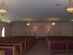 Rows of red pew seats in a chapel, ornate chandeliers, stage at far end.
