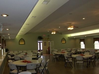 Empty event hall with round tables and white chairs. Soft lighting, light green walls, and decorative arched windows.