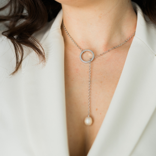 Woman wearing a silver necklace with a circular pendant and pearl drop, over a white blazer.