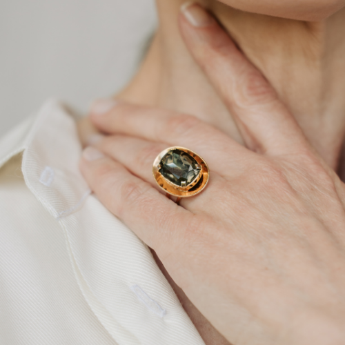 A hand wearing a gold ring with a large, oval, dark green gemstone, resting on a person’s neck.