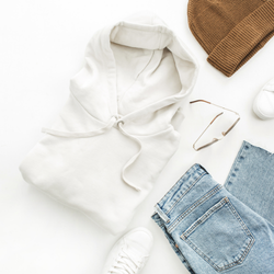 Cream-colored hoodie and white sneakers laid on a white surface with jeans, a brown beanie, and sunglasses.