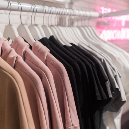 Clothes hanging on a rack; tan, pink, black, gray, and white shirts on white hangers against a white brick wall.