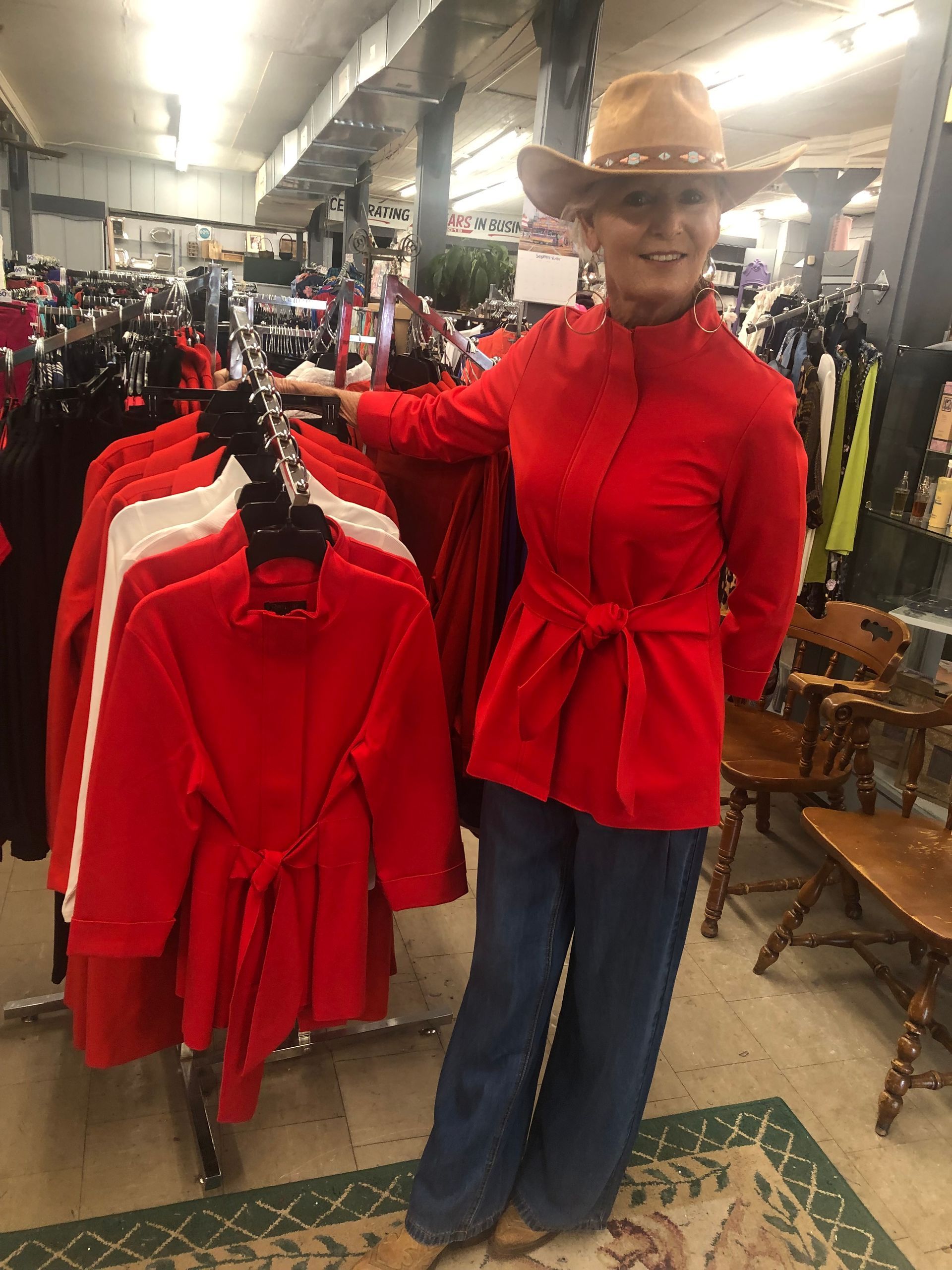 Woman in red jacket and cowboy hat stands by clothing rack with red jackets in a store.