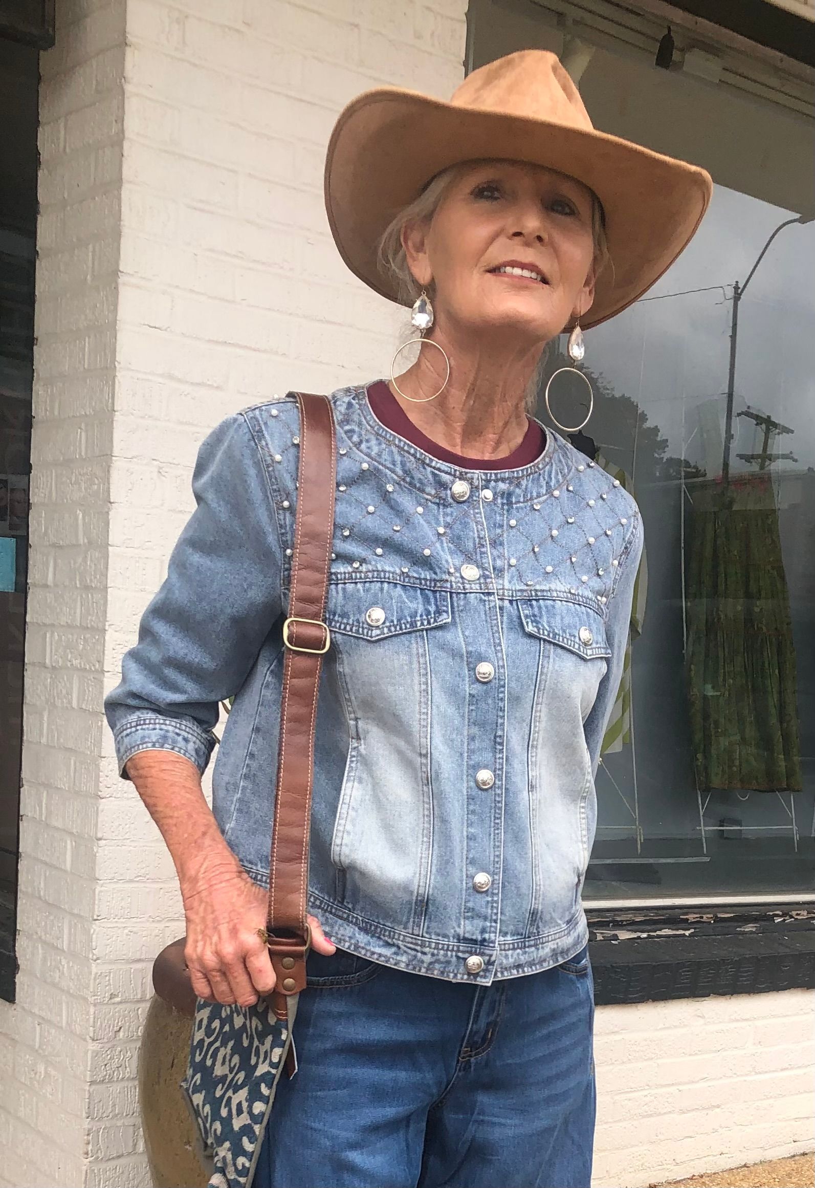 Woman in a denim jacket and cowboy hat, smiling and holding a bag, standing outside a brick building.