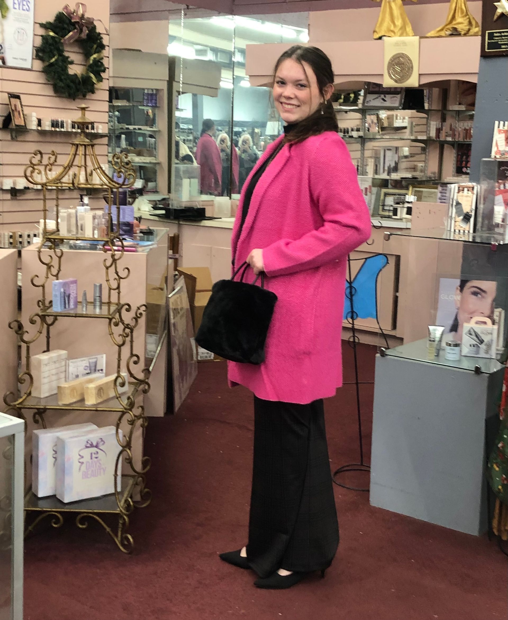 Woman in pink coat and black pants holds a black purse, standing in a shop with shelves.