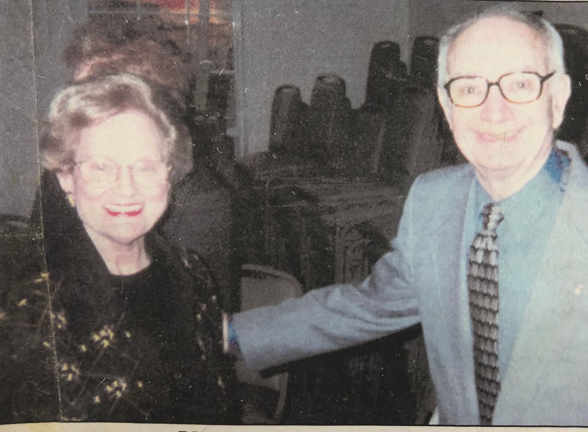 Older couple smiling; woman in black patterned top, man in blue suit and tie, standing indoors with chairs in the background.