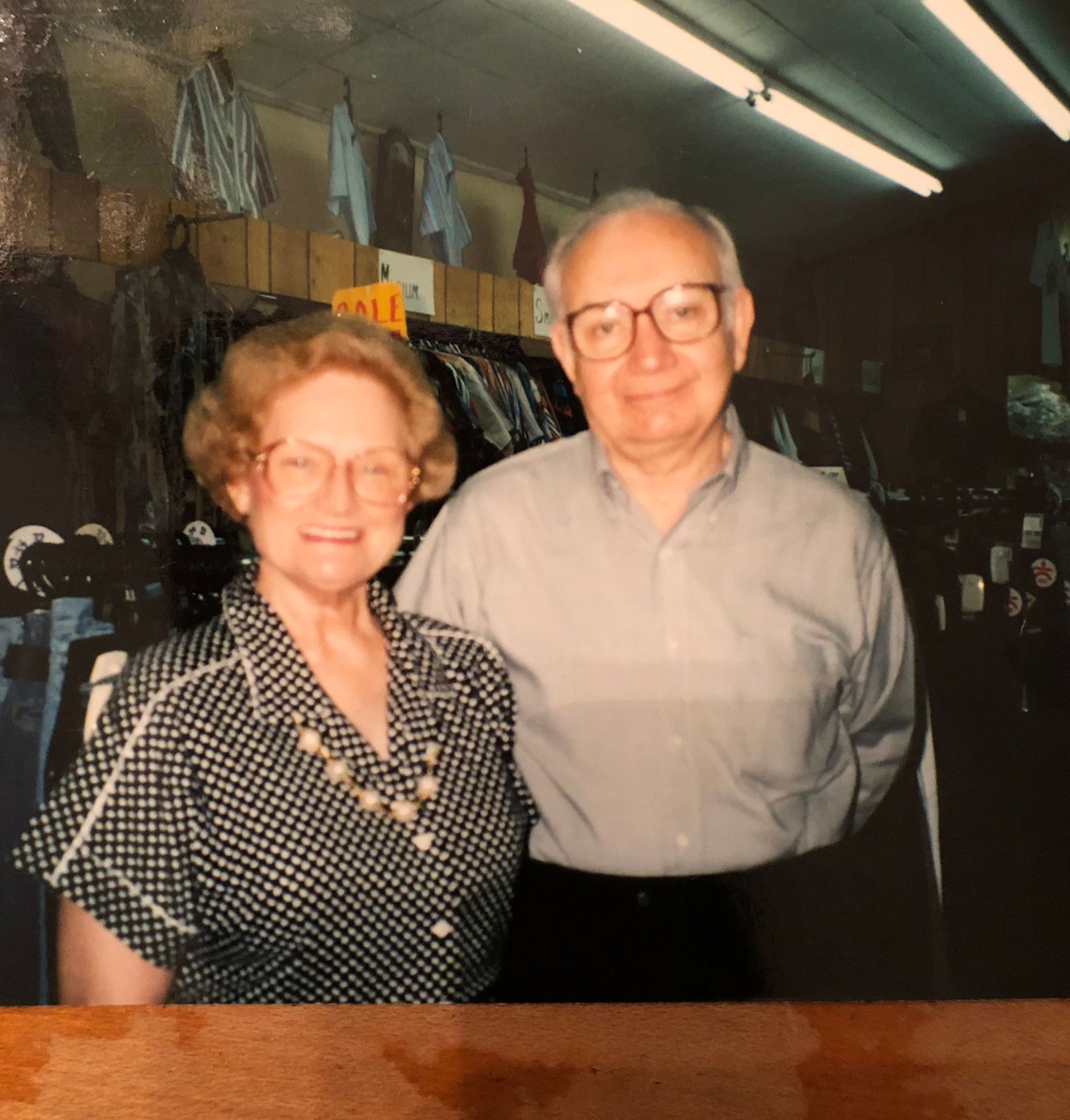 An older couple stands in a store. The woman in a patterned shirt smiles. The man wears a light shirt and glasses.