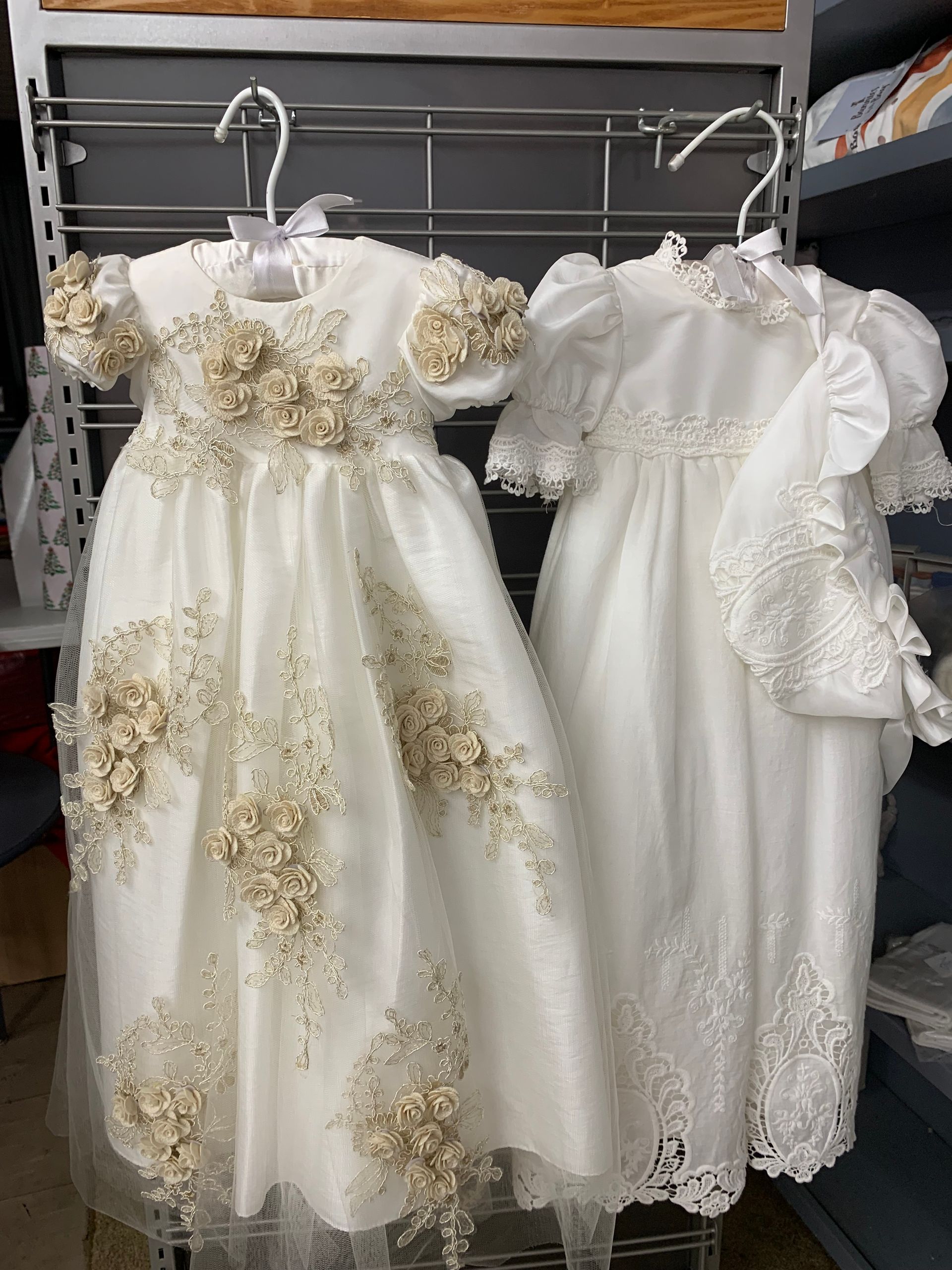 Two white baby dresses with floral and lace details hanging on metal racks.