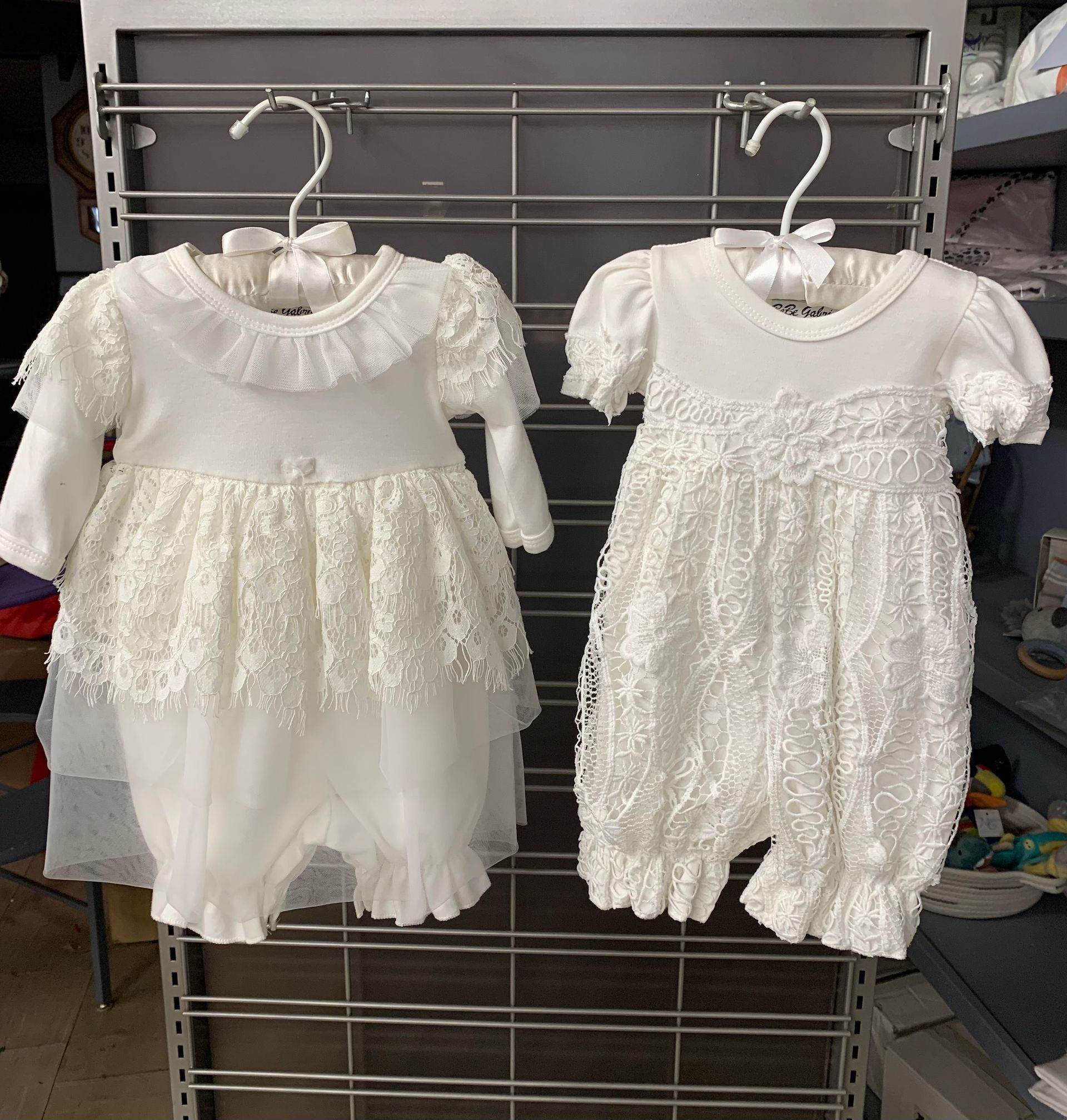 Two white lace baby rompers hanging on a metal rack.