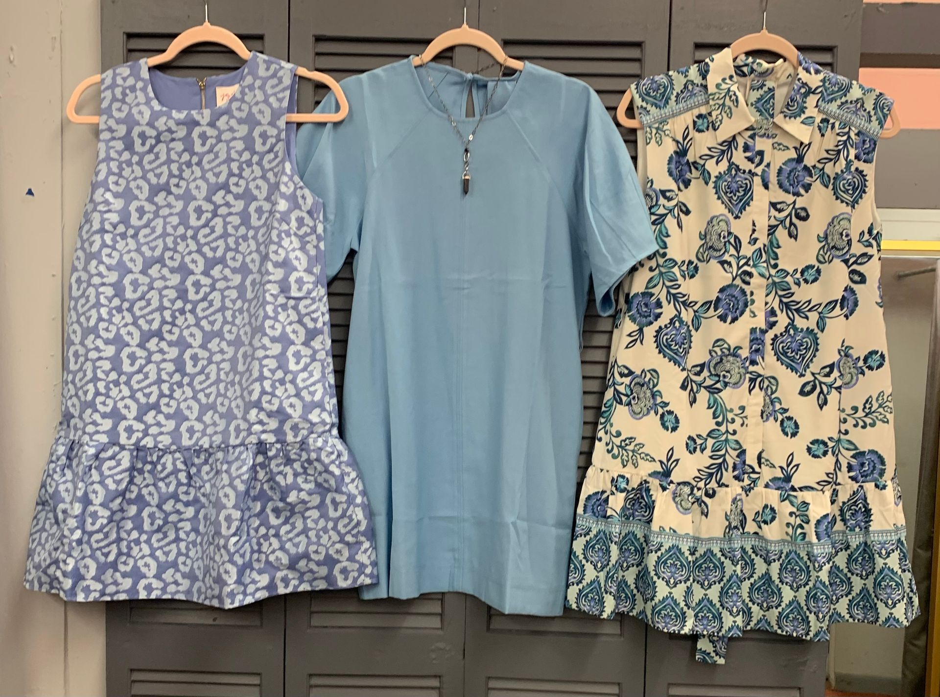 Three pastel dresses hanging on wooden hangers against shuttered doors.