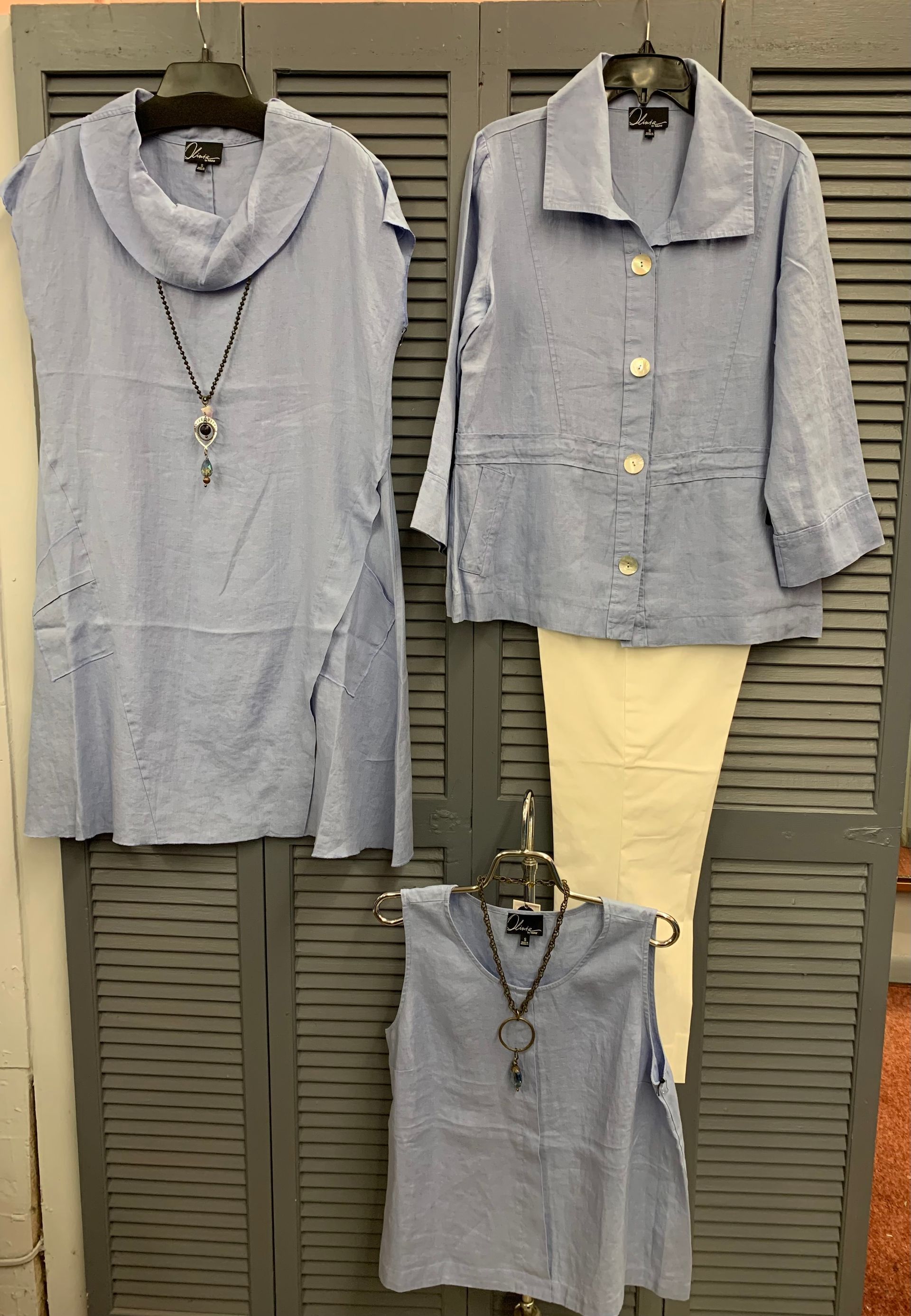 Blue linen clothing items, including tops and a jacket, displayed on hangers against a gray shutter background.