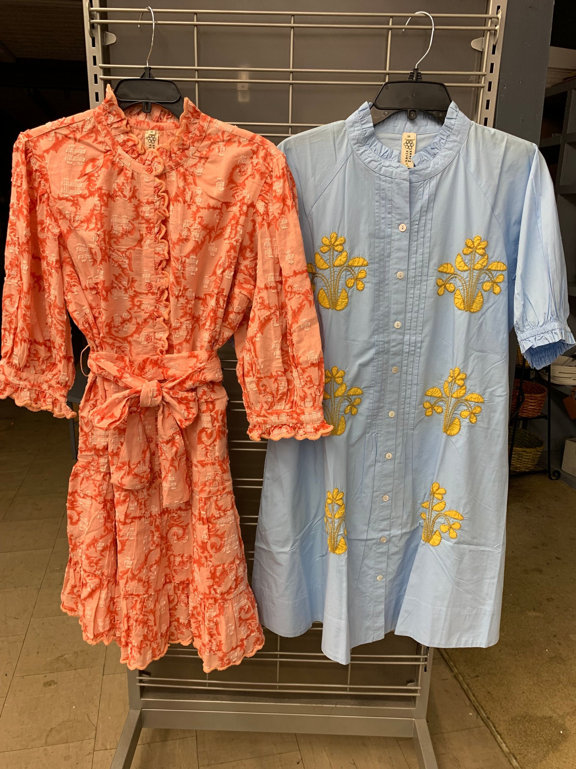 Two dresses on hangers: a ruffled peach floral dress with a sash and a light blue dress with yellow embroidery.