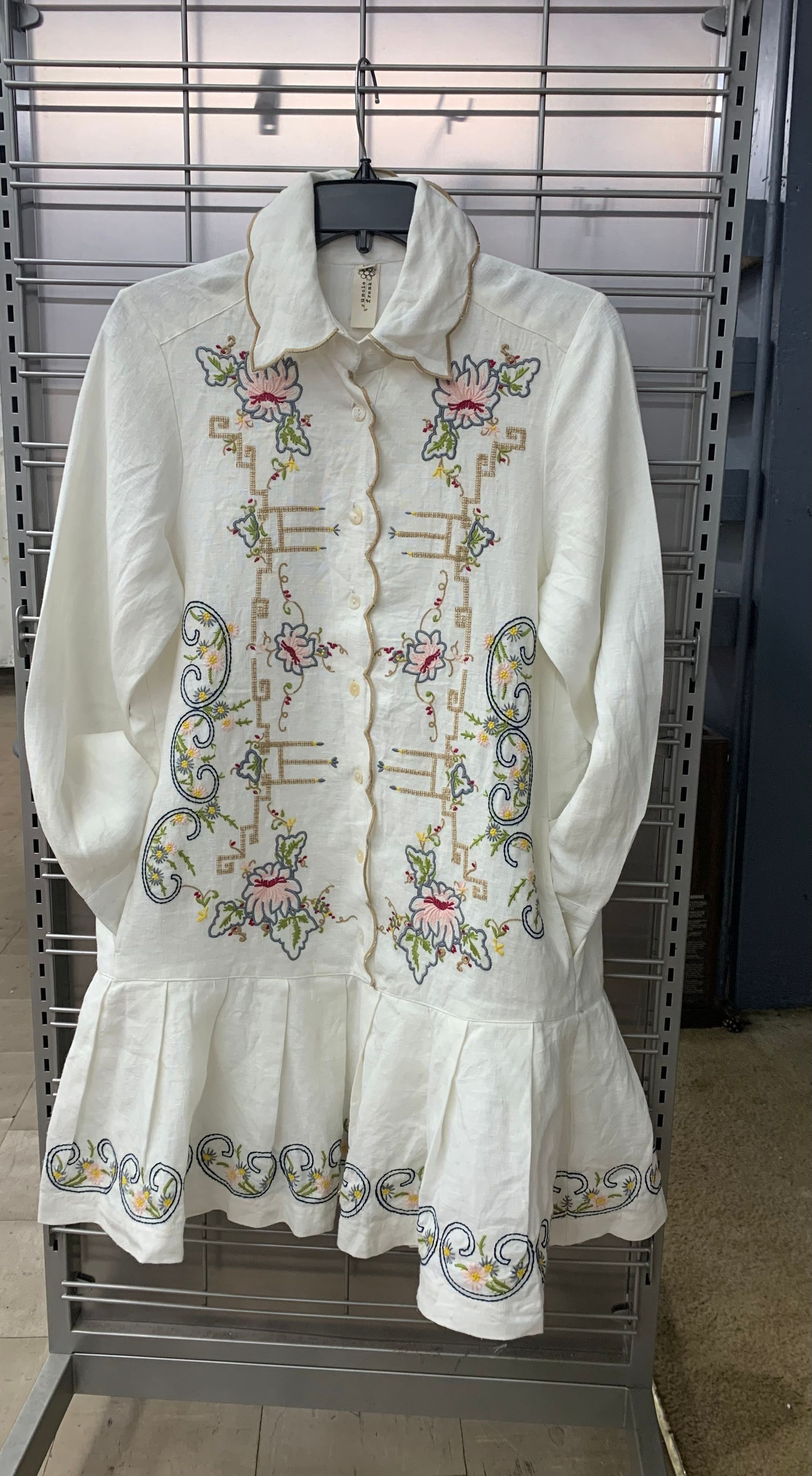 A white long-sleeved dress with button-down front, floral embroidery, and a ruffled hem, hanging on a wire rack.