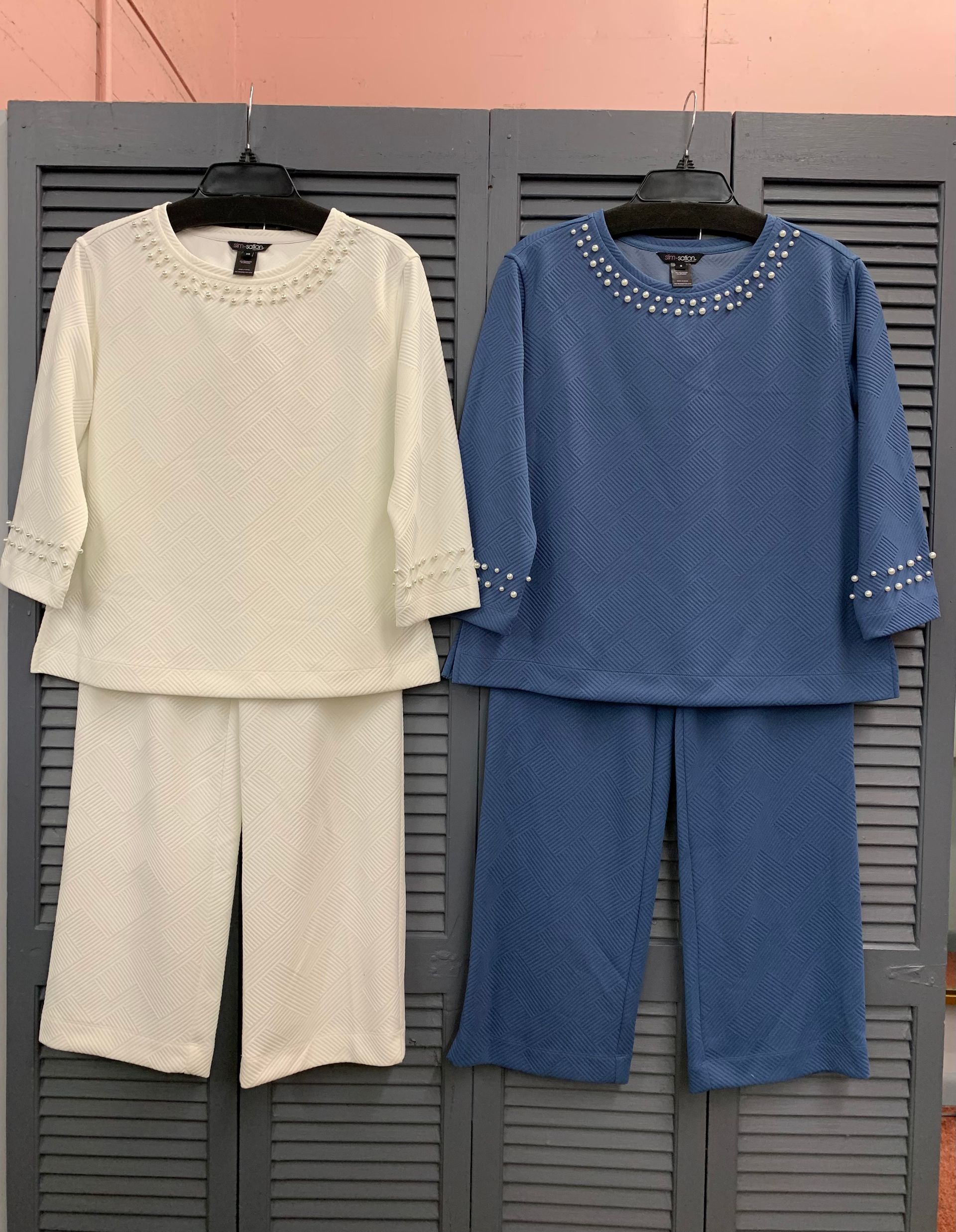 Person seated on a wooden stool, wearing a cap, grey sweater, jeans, and sneakers. Leaning on arm, looking down.