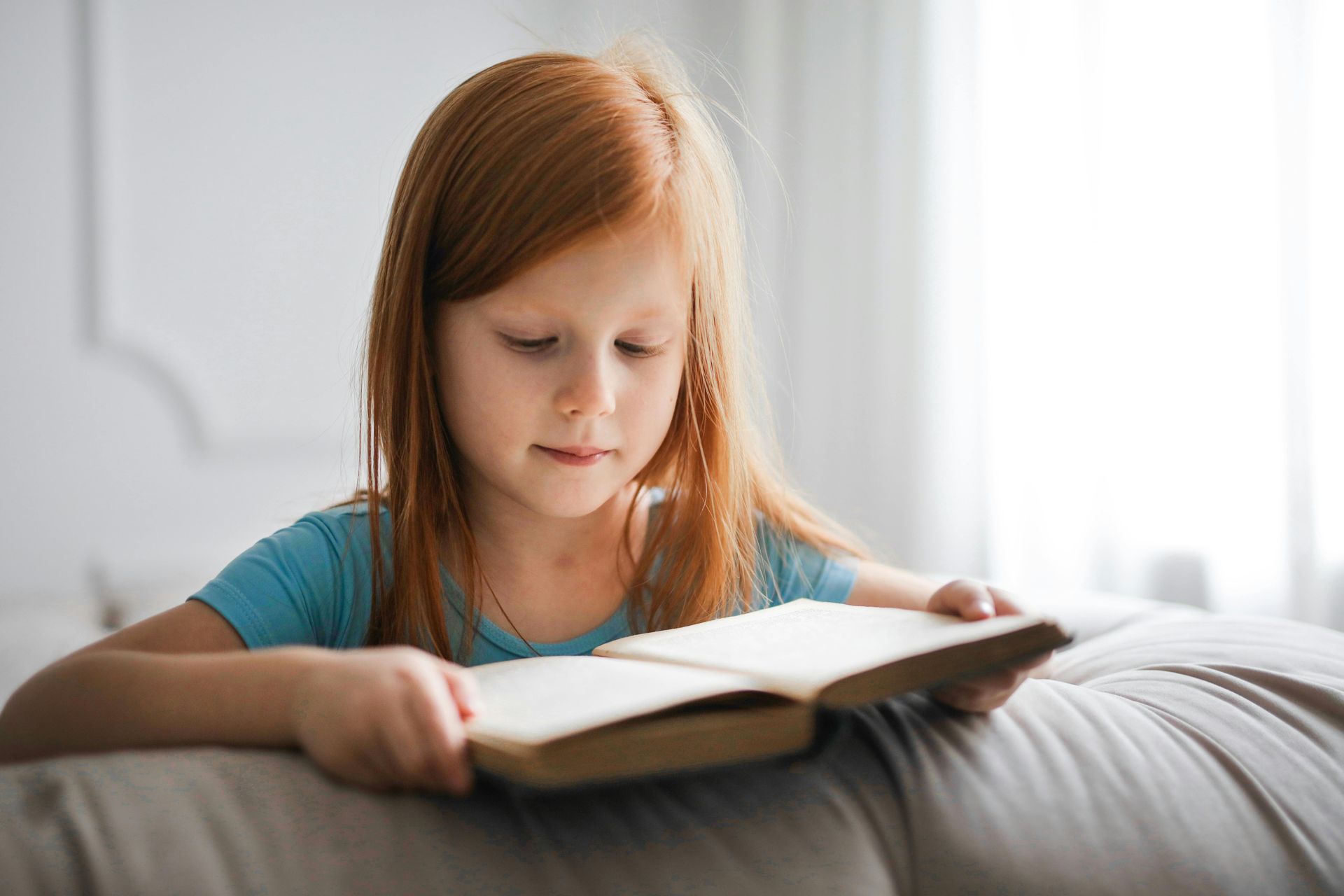 Young girl with red hair reading a book on a couch.
