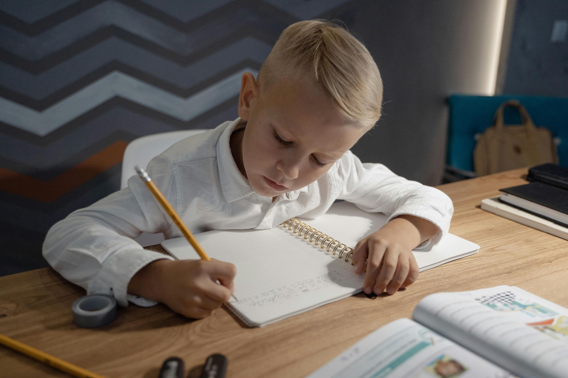 Boy with blonde hair, writing in a notebook at a wooden desk.
