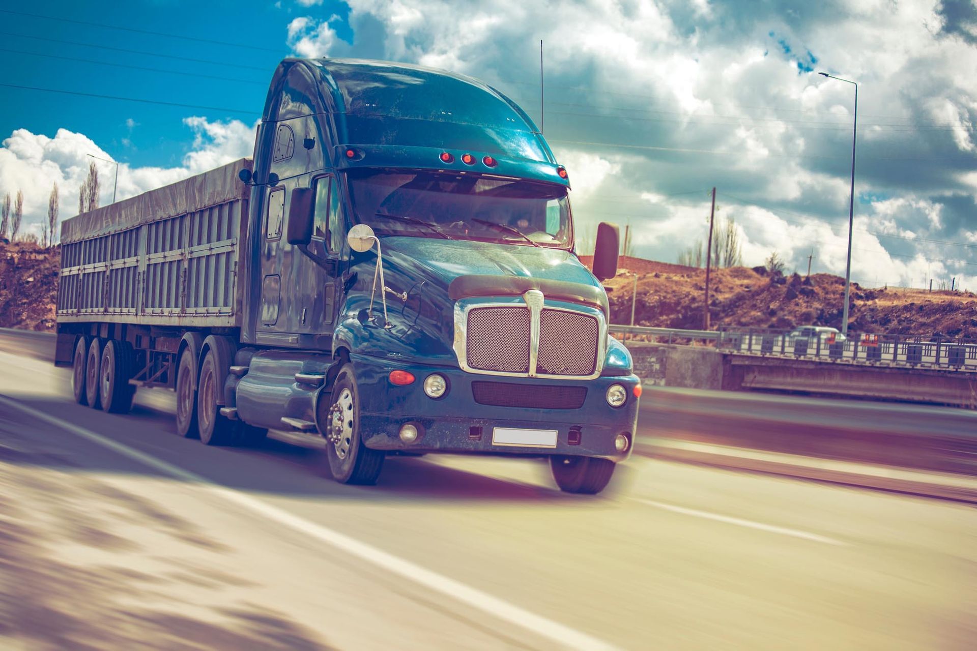 Dark blue semi-truck driving on a highway with an empty trailer, sunny sky in the background.