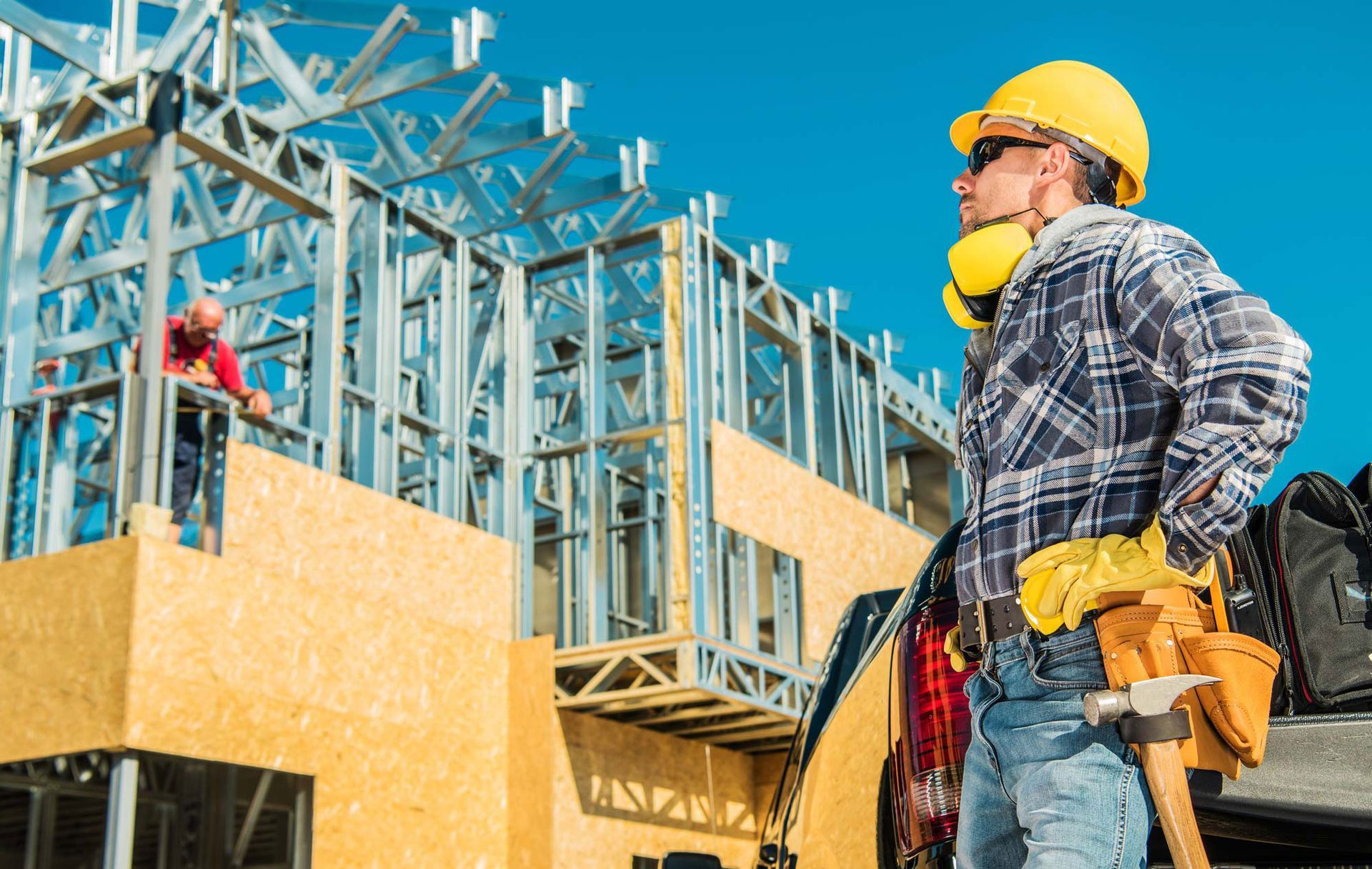 Construction worker wearing safety gear at a building site, looking upward. Another worker building in background.