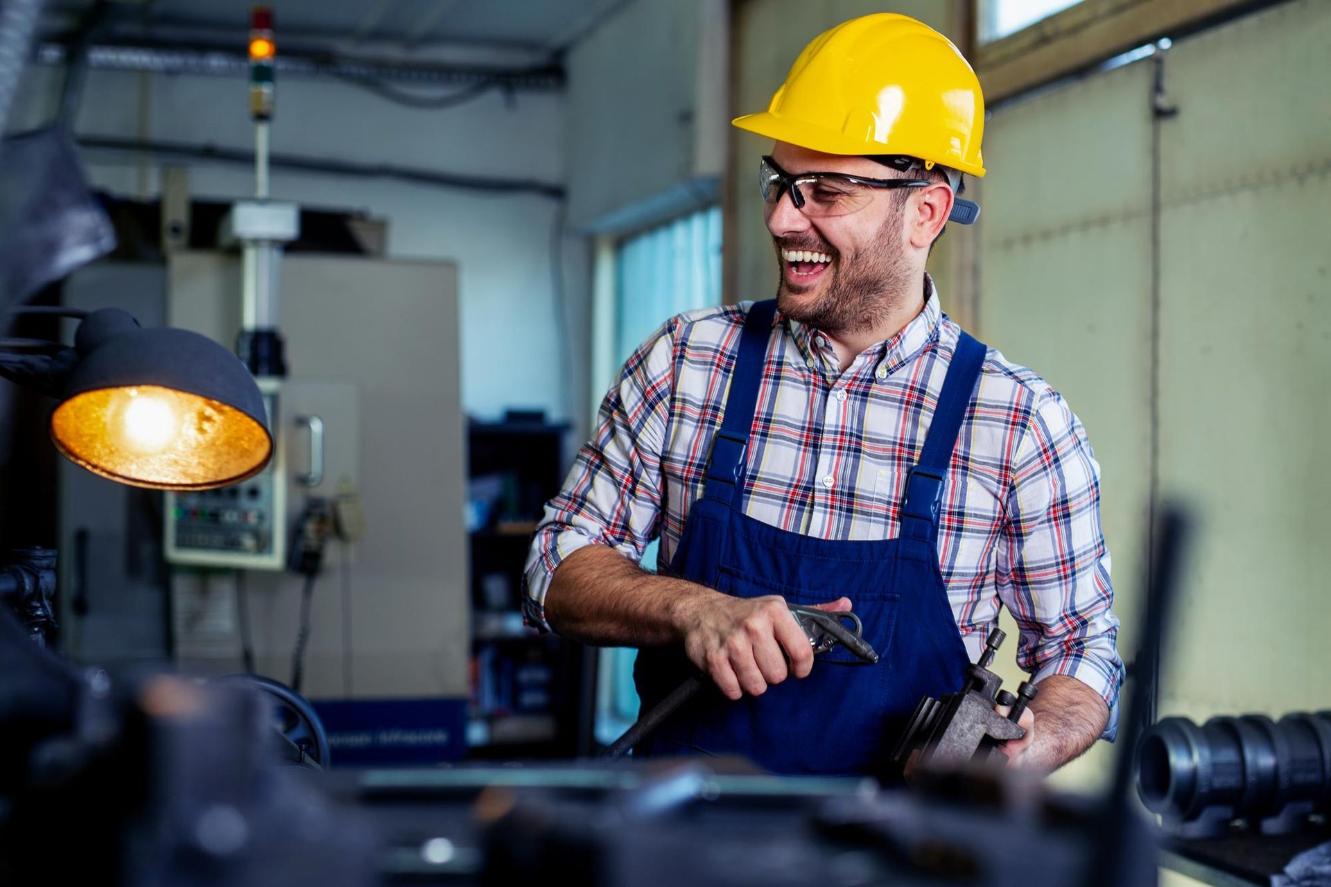 Man in yellow hard hat and overalls laughs in a workshop, holding tools.