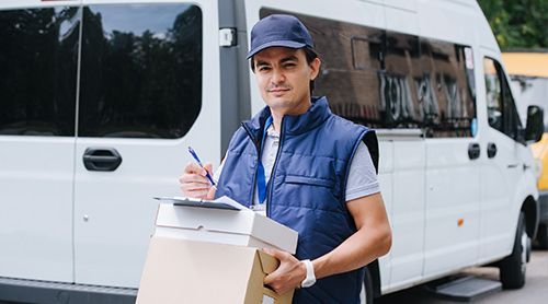 A delivery man is holding a box and a clipboard in front of a white van.