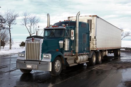 Semi-truck with teal cab and white trailer, parked on wet asphalt next to a snowy landscape.