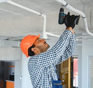 A worker in an orange hard hat and plaid shirt uses a power drill to install a pipe on a concrete ceiling.