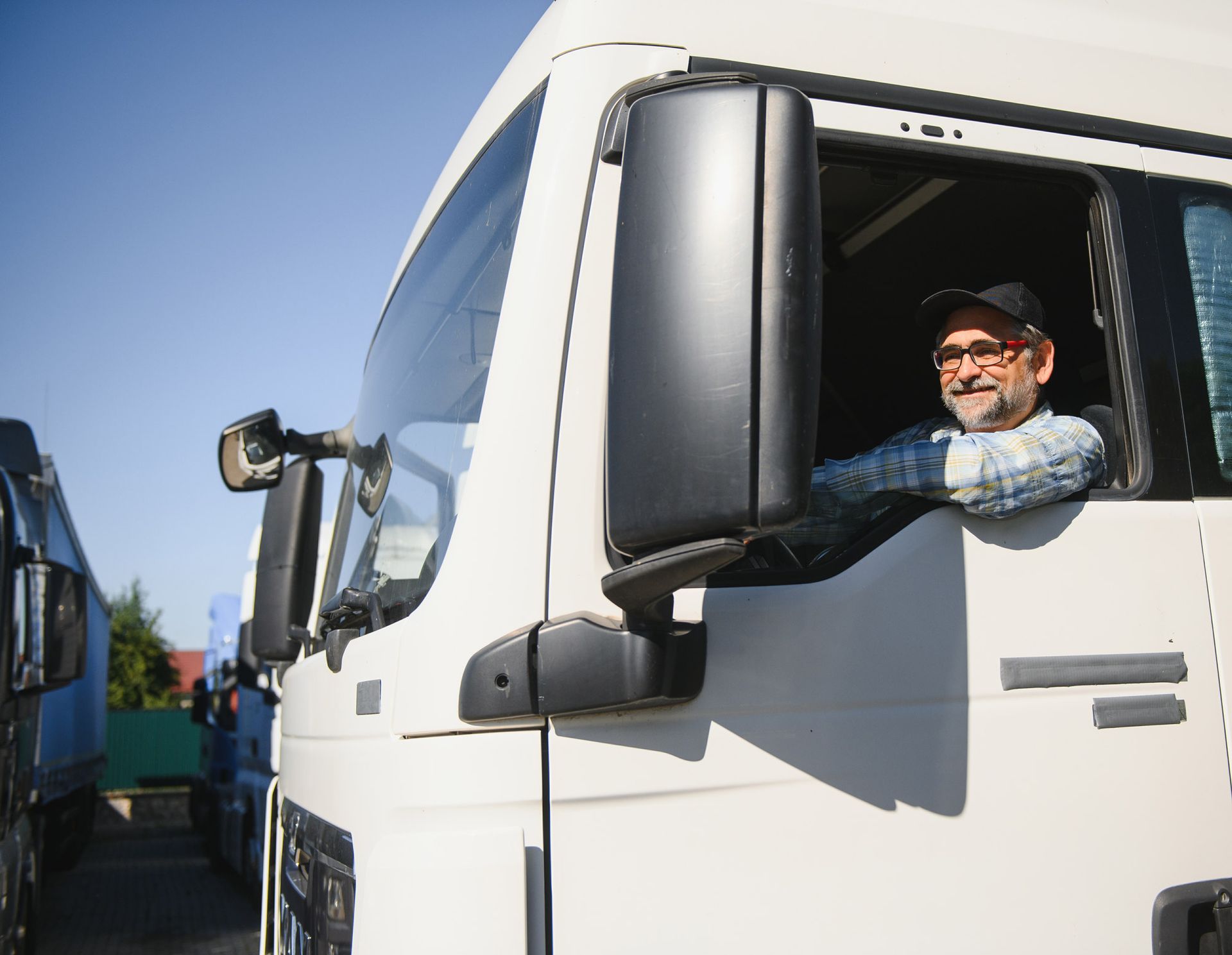 Truck driver with a beard and glasses smiling from the cabin of his white truck on a sunny day.