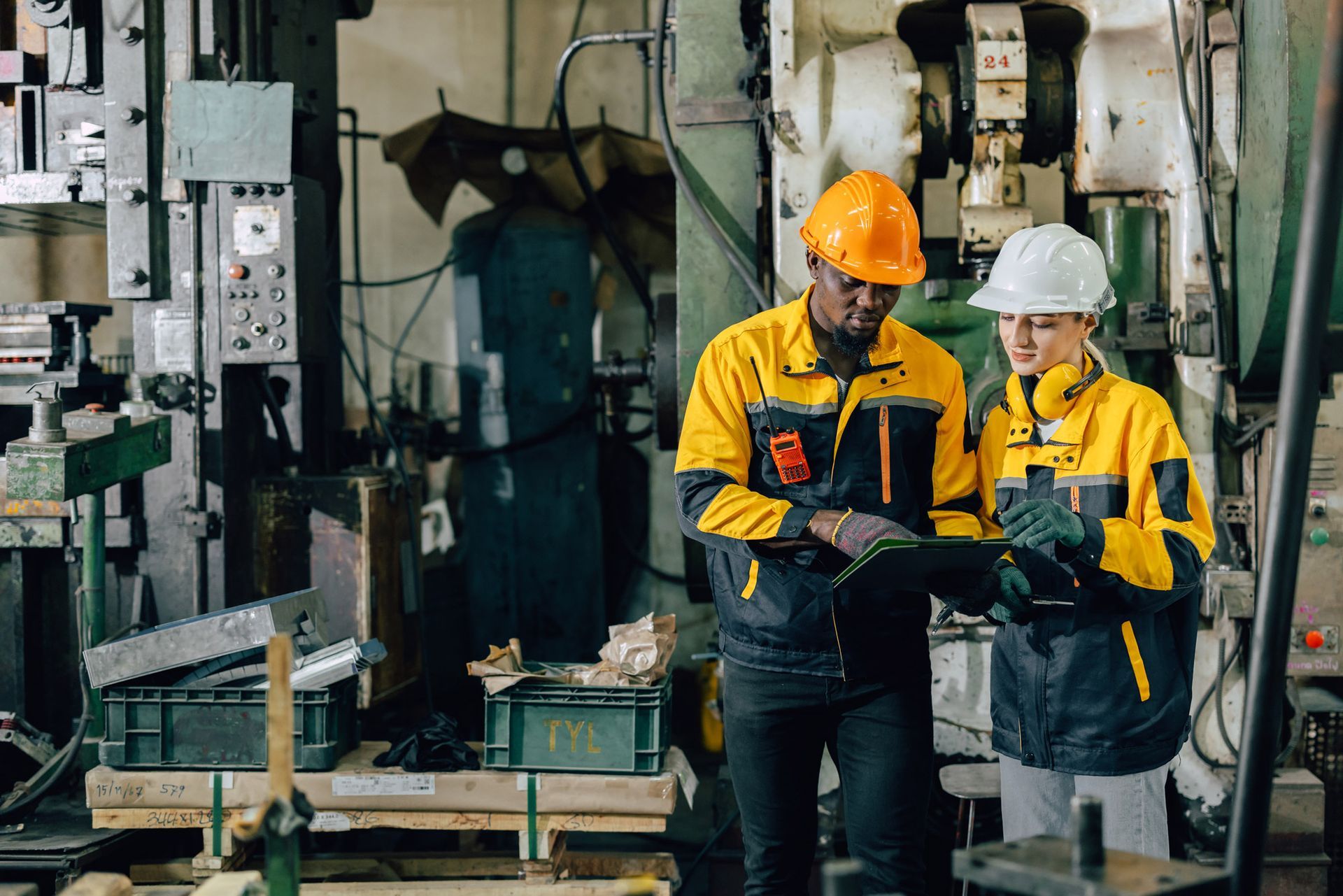 Two factory workers in safety gear reviewing a tablet near machinery.