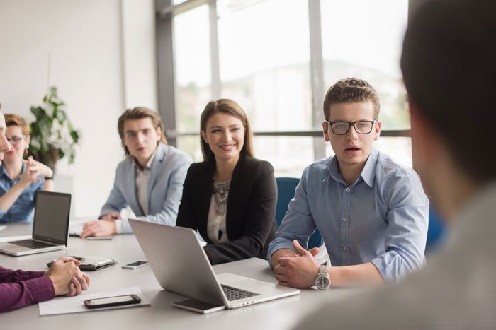 A group of diverse people in a meeting, using laptops.  Smiling woman in blazer, man with glasses talking.