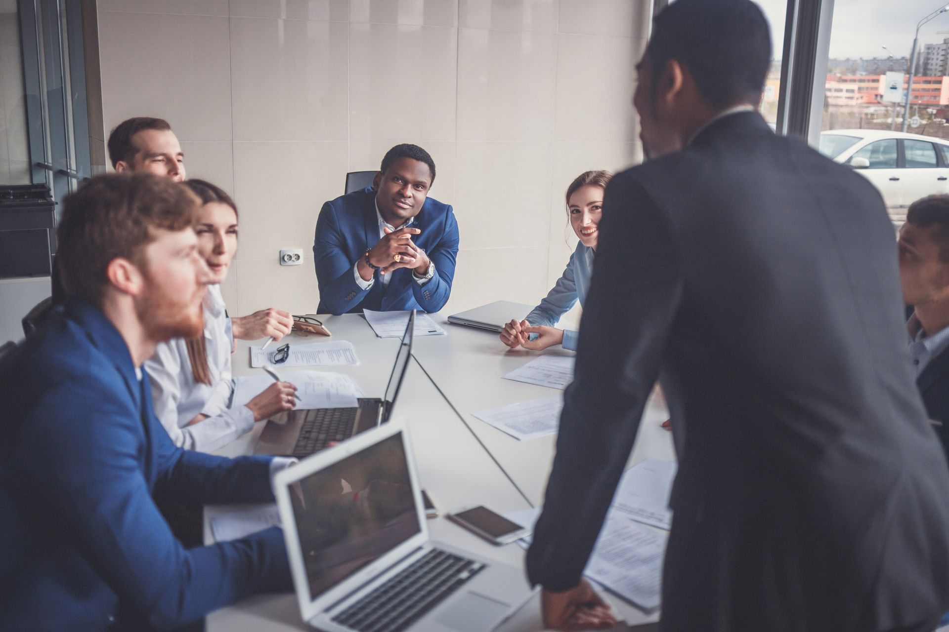 Business meeting: diverse group around a table, focused, discussing, led by a person standing and presenting.