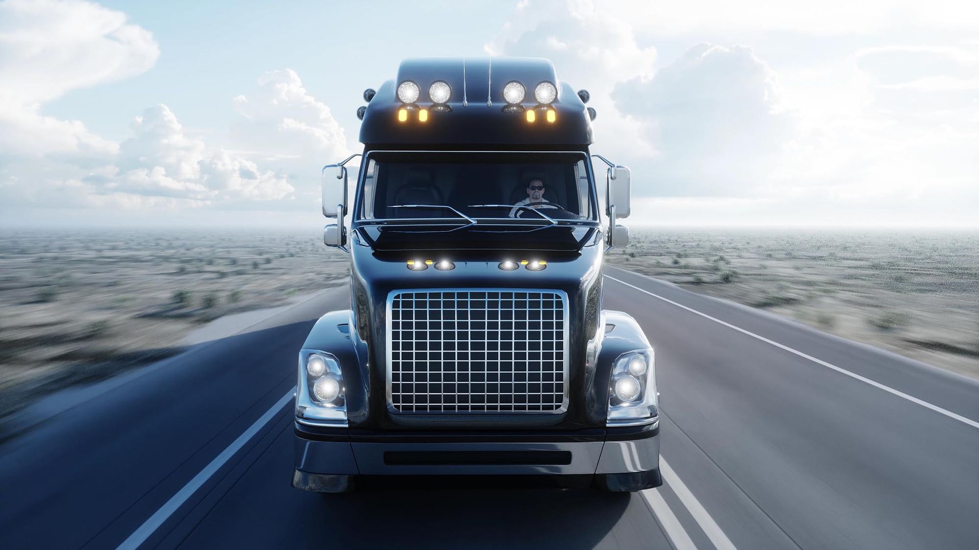 Black semi-truck driving towards the viewer on a highway under a bright blue sky.