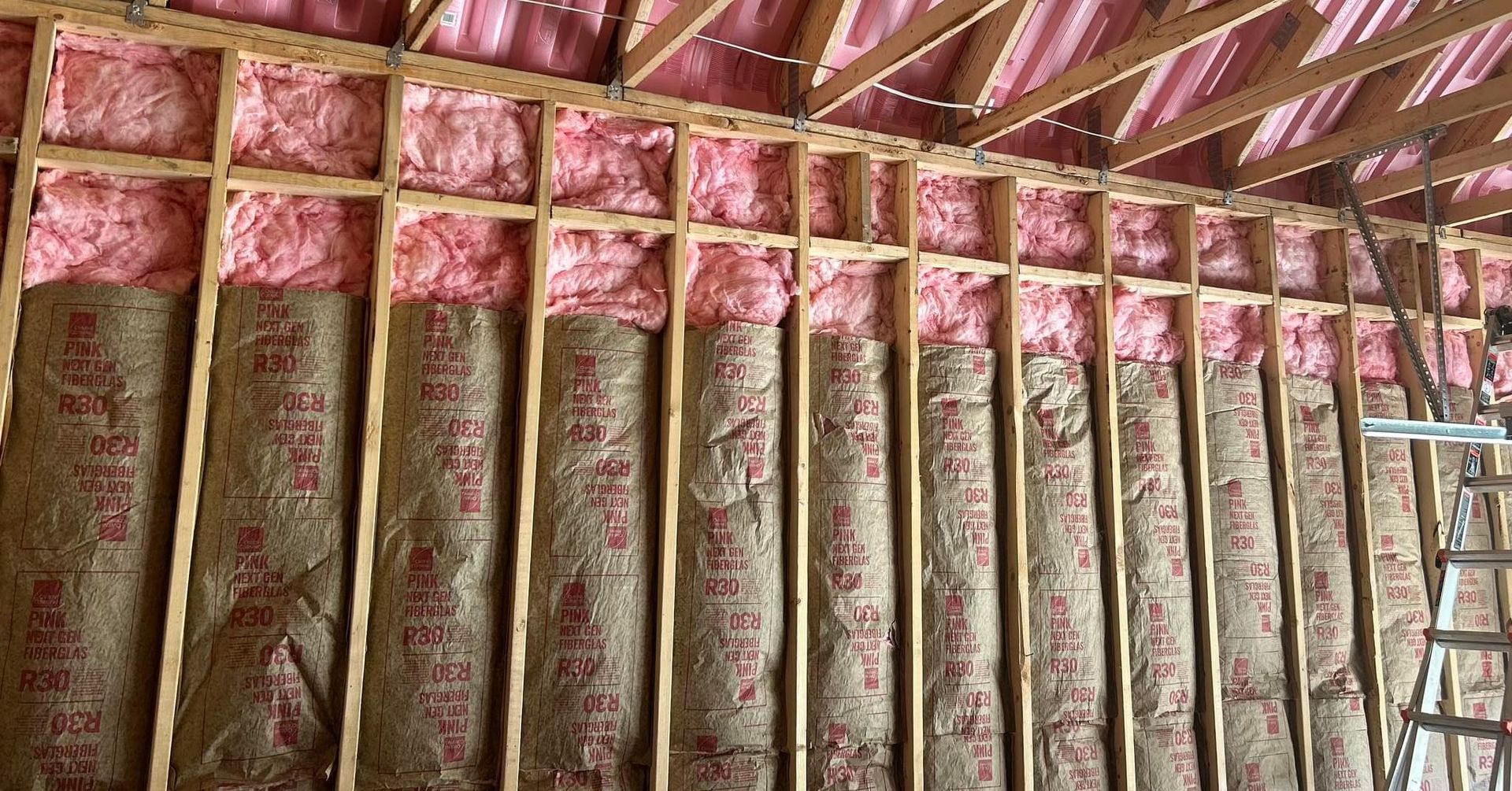 Pink insulation rolls installed between wooden wall studs in a construction site.