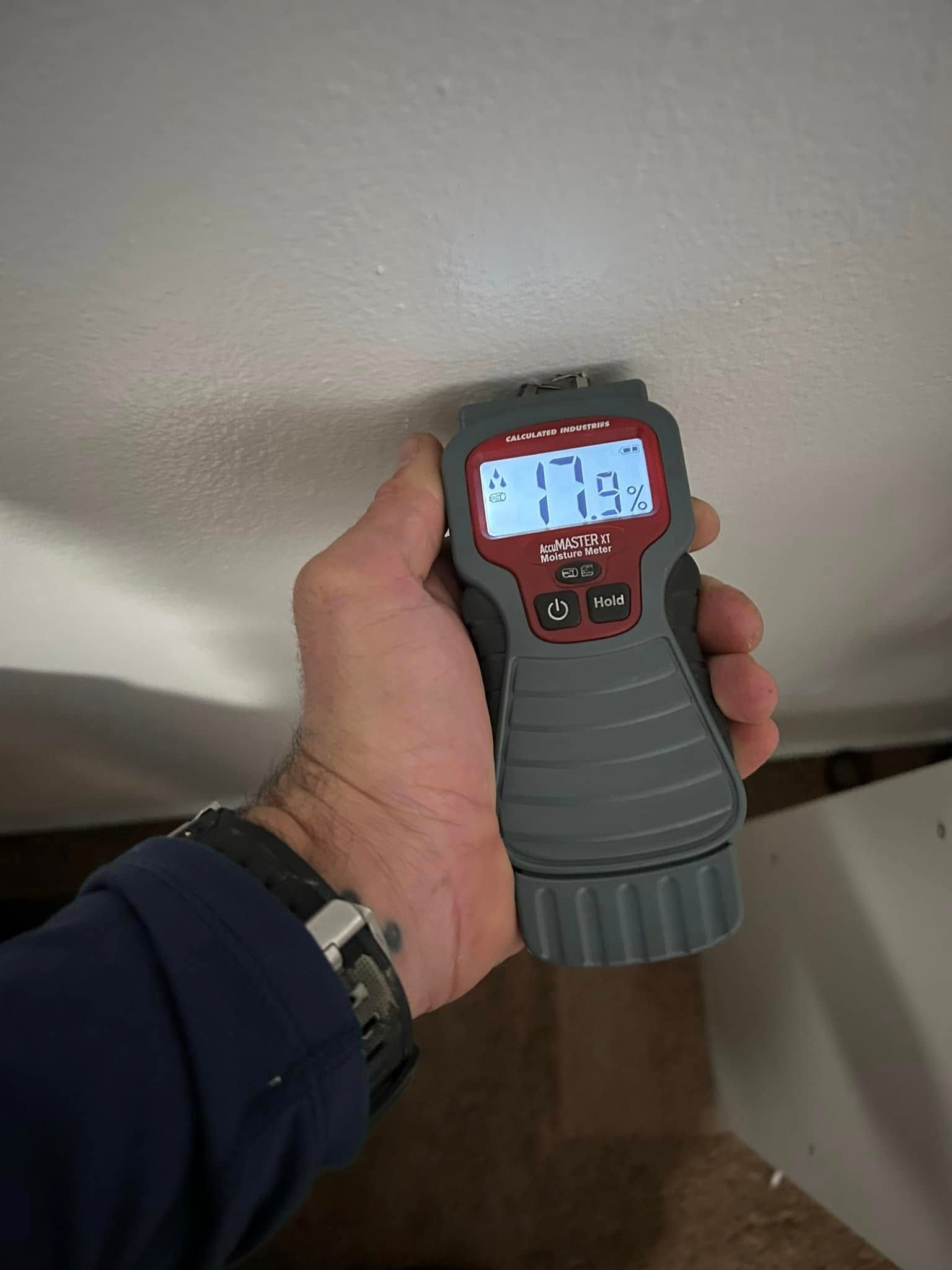 Hand holding moisture meter, reading 17.9% on a white ceiling, possibly checking for water damage.