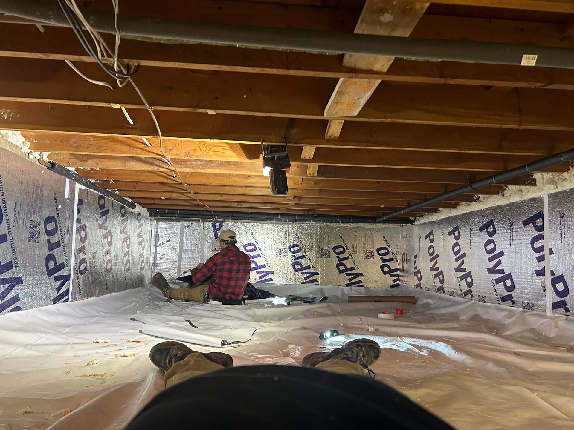 Person inside crawl space, installing insulation. Silver foil insulation lines the walls, wood beams above.