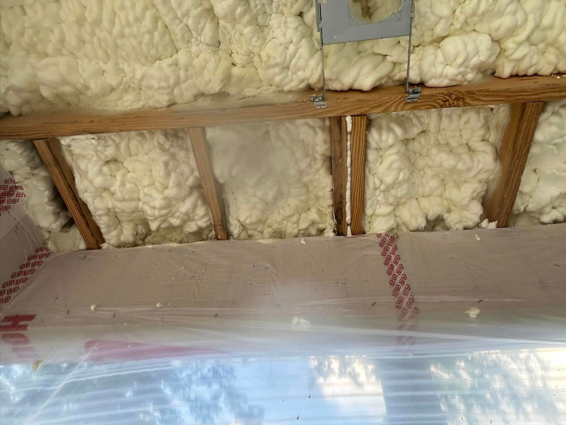 White spray foam insulation in a wooden attic, covered by clear plastic and tape.