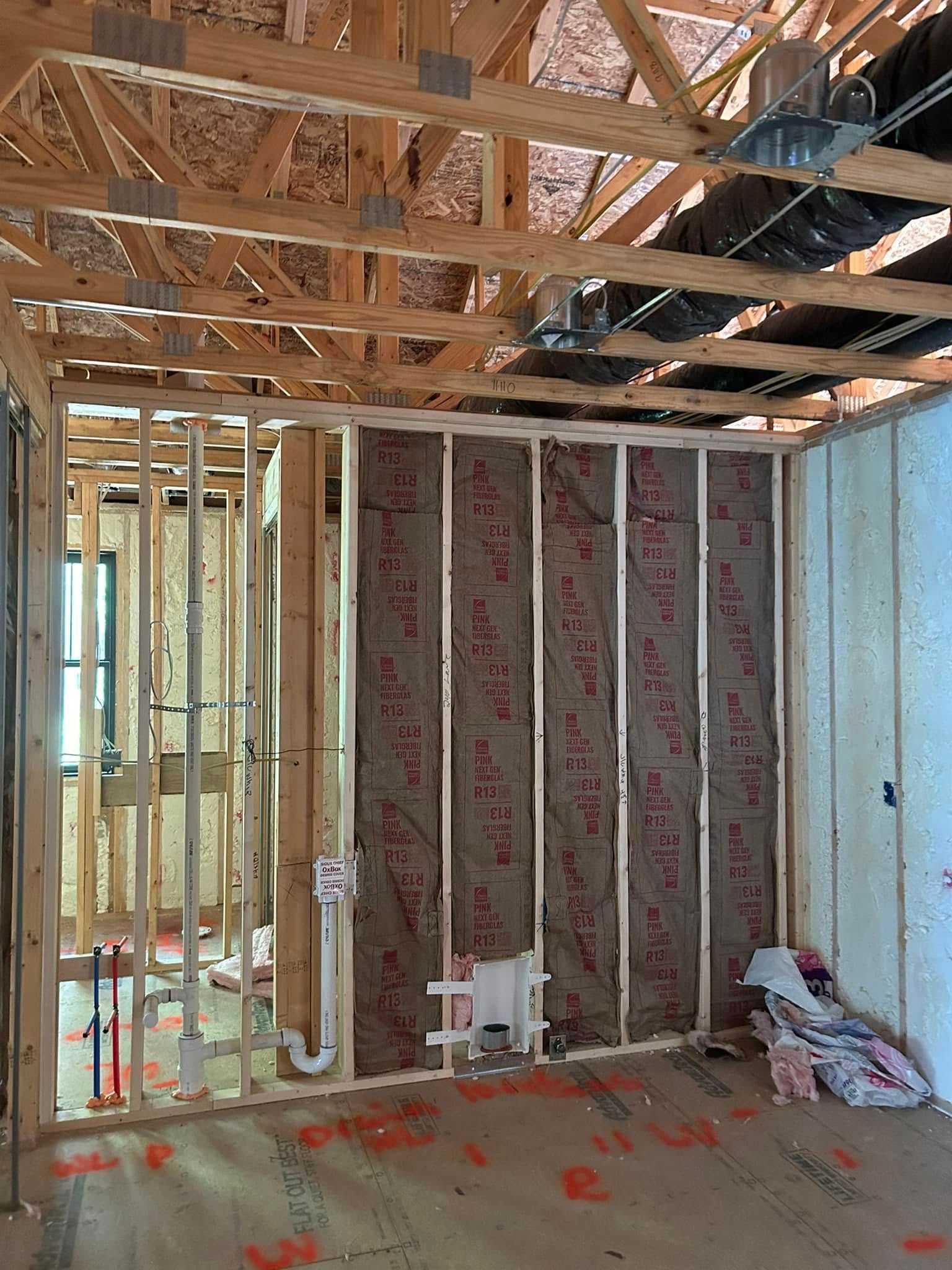 Interior view of a house under construction; wood framing, insulation, and plumbing visible.