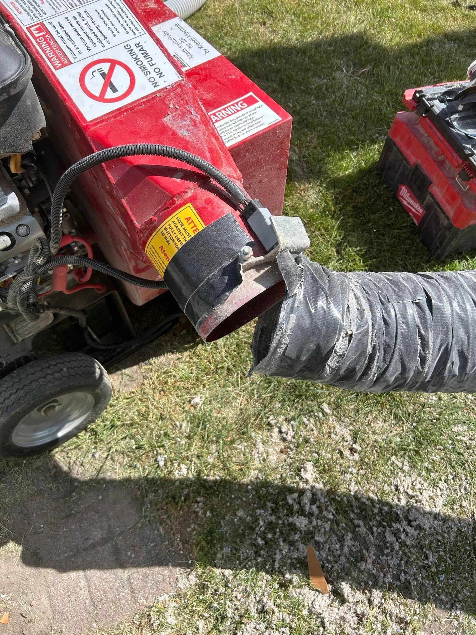 A red leaf blower with a black hose sits on grass next to a sidewalk.