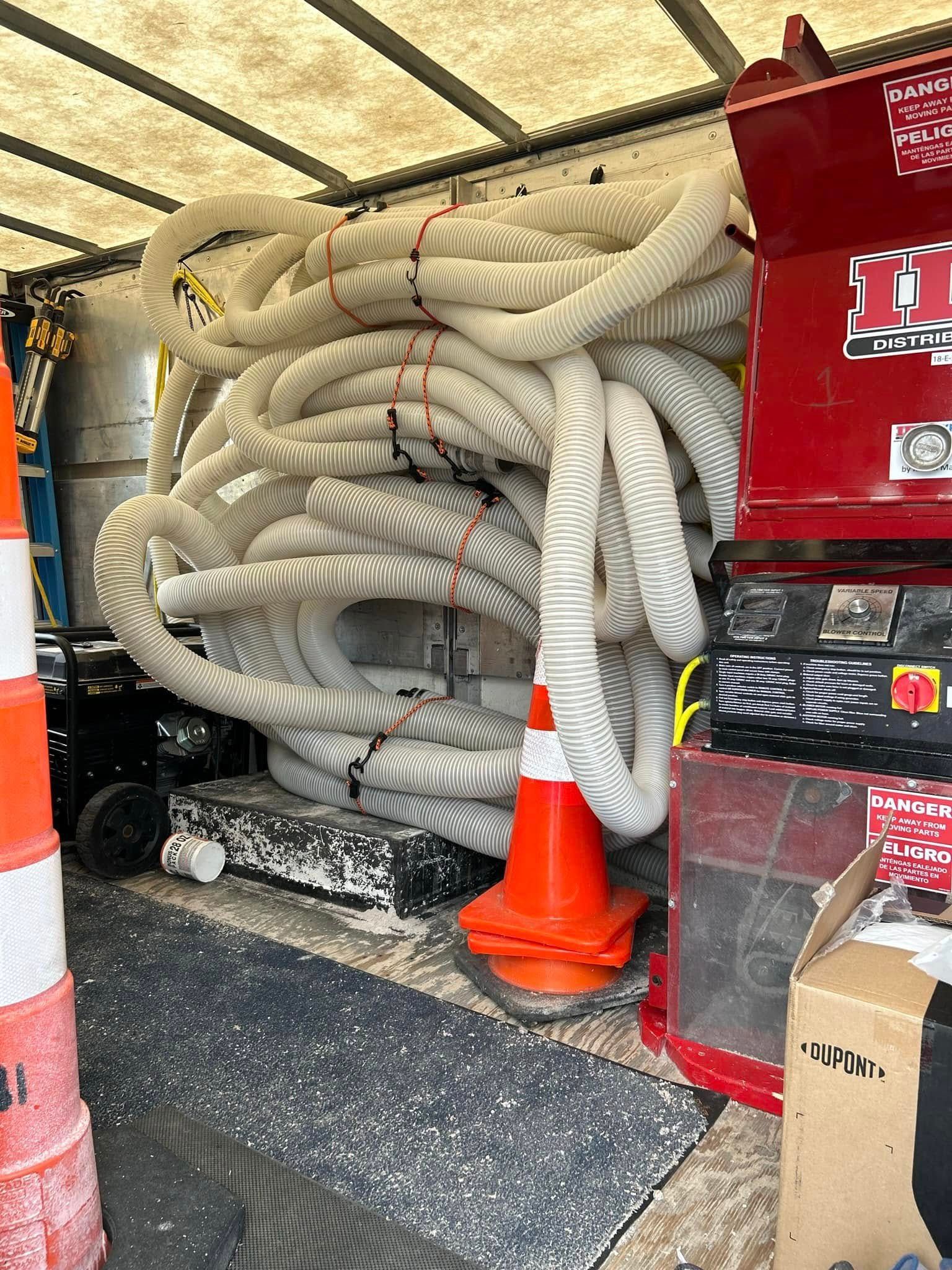 Inside a truck, white hoses are bundled with zip ties next to a red machine, traffic cones, and other equipment.