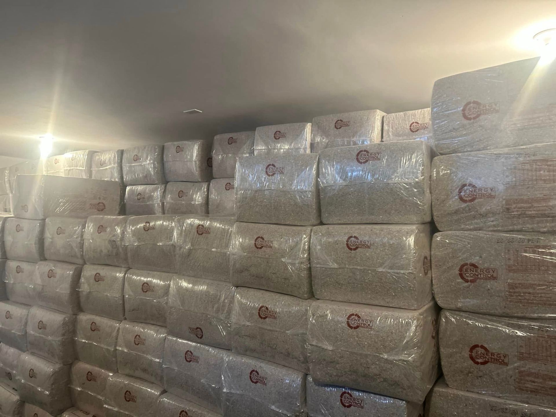 Stacks of packaged goods fill a storage room. The packages are wrapped in clear plastic with a circular logo.