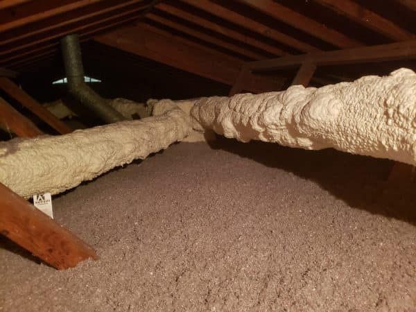 Attic interior with grey insulation and a duct covered in white foam. Wooden beams are visible.