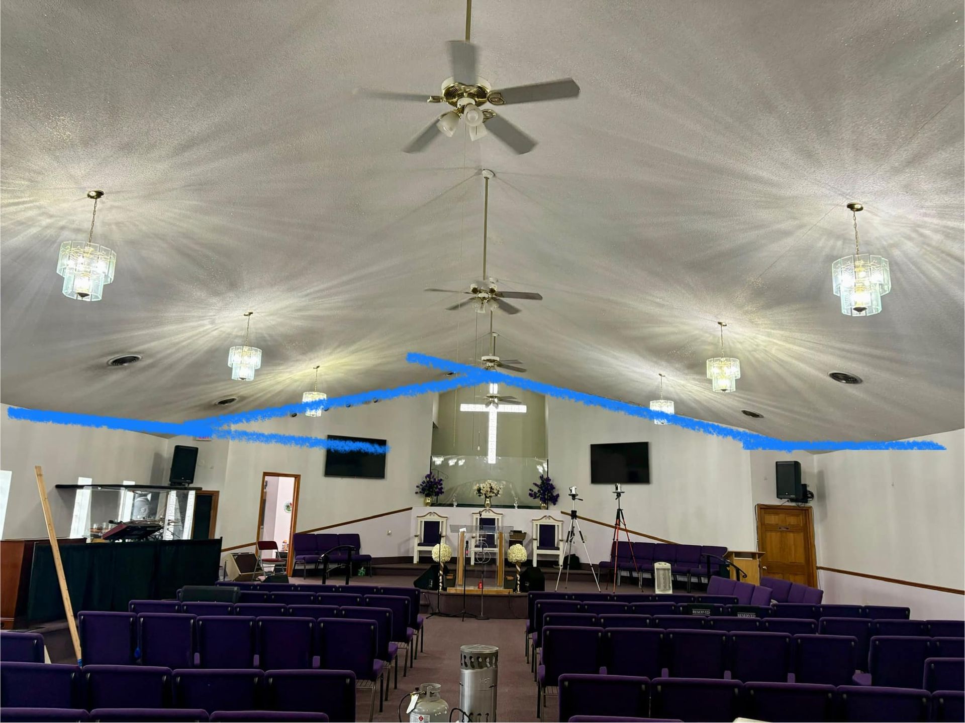 Interior of a church with rows of purple pews, a stage with a cross, and a high ceiling with chandeliers.