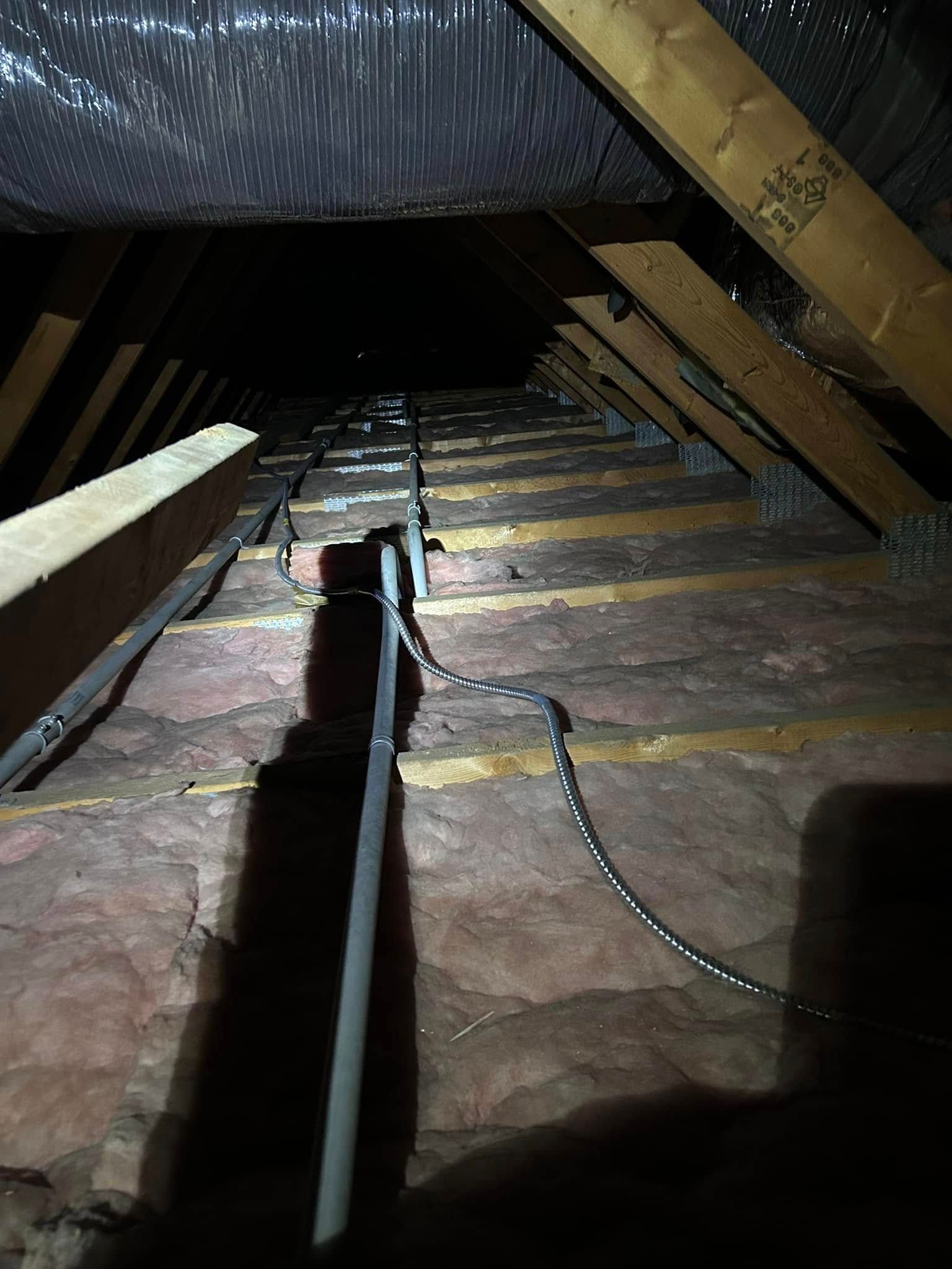 Attic view: Insulation, wooden rafters, electrical wiring, and a ladder in a dark, cramped space.